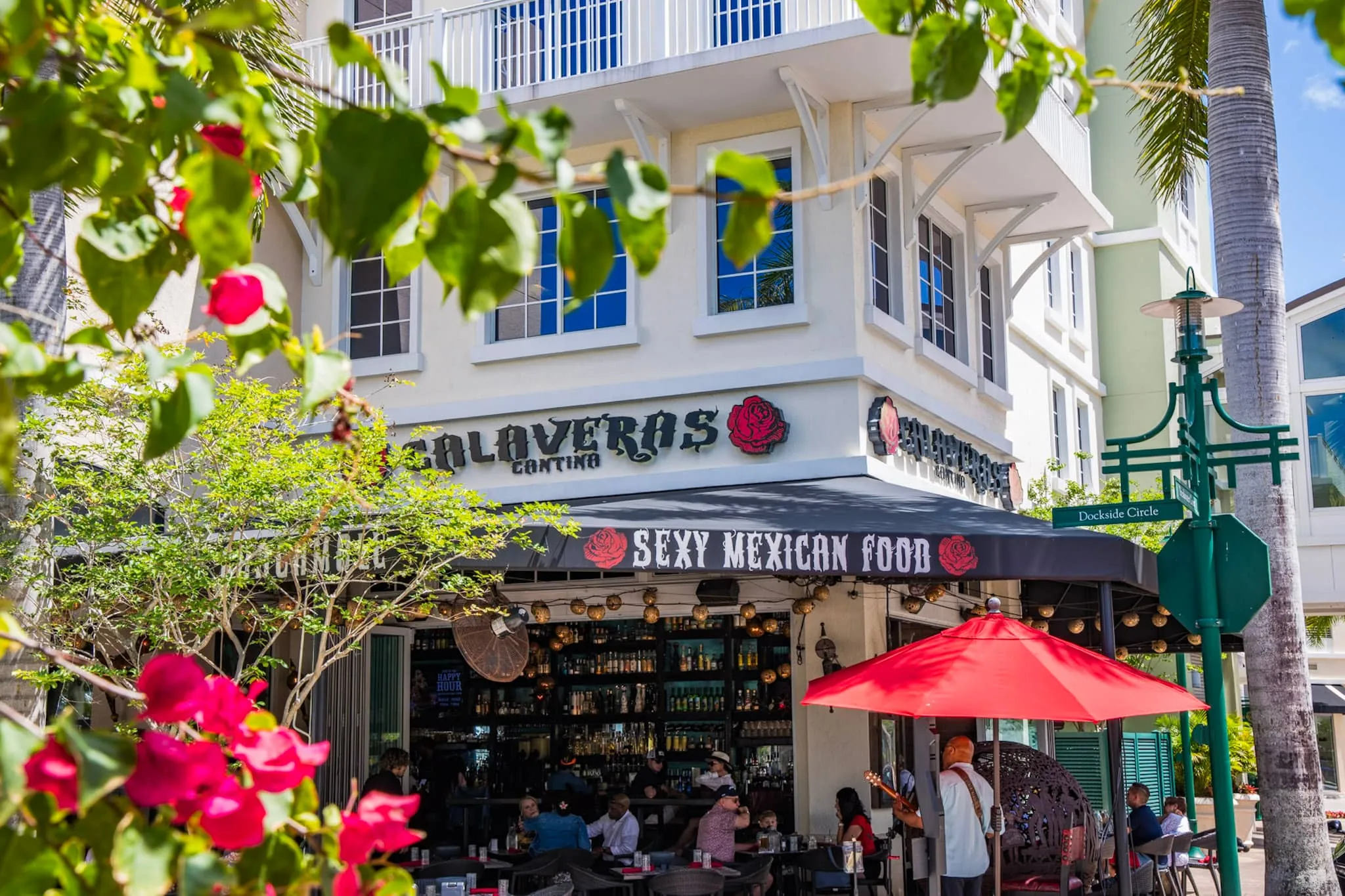 Cantina exterior with sidewalk seating and a red market umbrella under bougainvillea